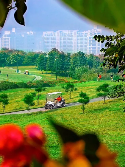 A photograph of the DLF Golf Course, framed by foliage to create depth. This image captures the lush landscape against the backdrop of the city skyline, promoting a luxury lifestyle.