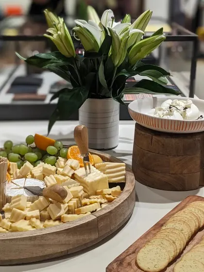 A beautifully styled cheese board for the Paul Smith event, featuring a wheel of Brie, cubed cheeses, fresh fruits, and crackers, complemented by a vase of white lilies.