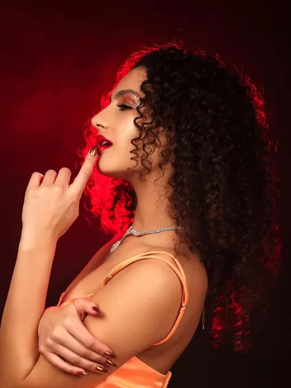 A beauty portrait that uses a red light to cast a warm glow, highlighting the model's features and curly hair. The pose is soft yet confident, creating a captivating mood.