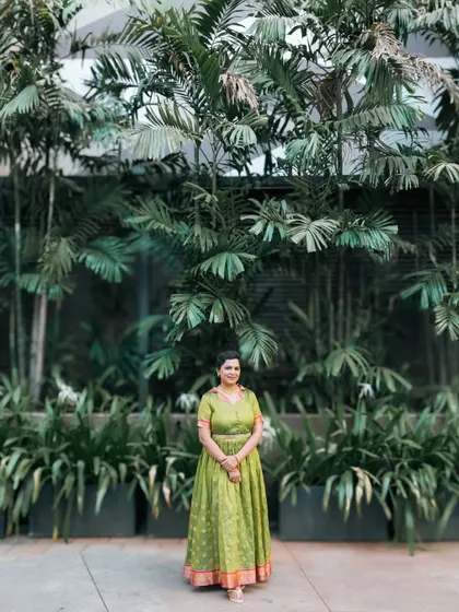 A wide shot of a woman in a traditional green dress against a wall of lush greenery.