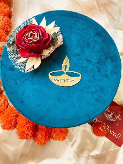 A luxurious round velvet box in a rich teal color, adorned with a floral accent and a golden 'Happy Diwali' emblem. This box is perfect for premium gifting.
