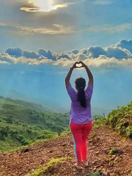 A trekker making a heart shape with her hands against the backdrop of the beautiful Chikmagalur hills.