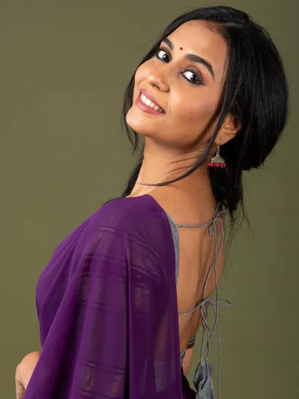 A smiling portrait that captures the simple beauty of the purple saree and the modern, deep-back blouse design.