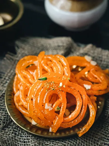 Hot, crispy jalebis. The moody lighting and rustic texture of the burlap underneath make the vibrant orange of the jalebis pop, emphasizing their sugary, crunchy appeal.
