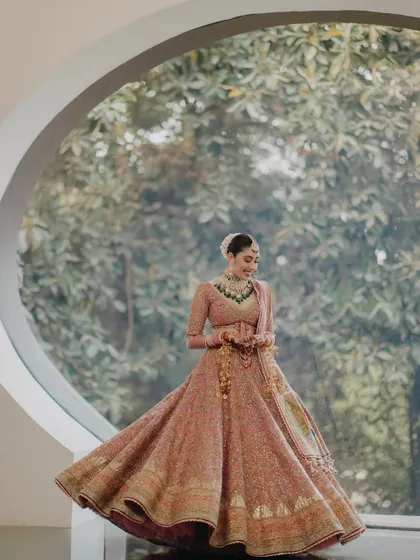 A stunning portrait of Ragini framed by a large circular window. The natural light and the lush green background create a beautiful contrast with her pink lehenga, resulting in a breathtaking shot.