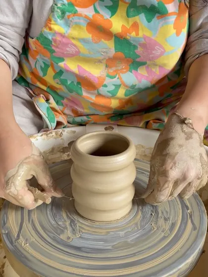 A student's hands covered in clay as she works on a piece at the wheel, capturing the tactile nature of pottery.