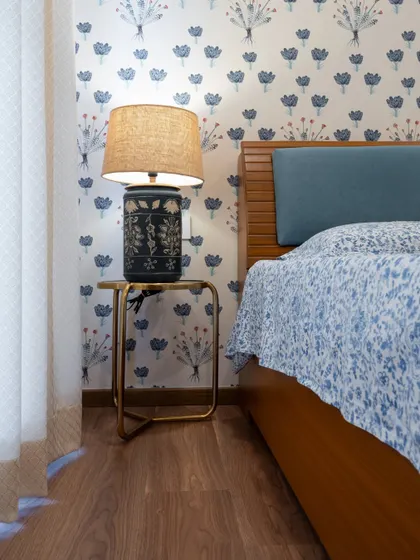 This bedroom has a charming, traditional Indian feel, with a floral block-print wallpaper and a classic wooden bed. The ceramic-base lamp adds to the ethnic aesthetic.