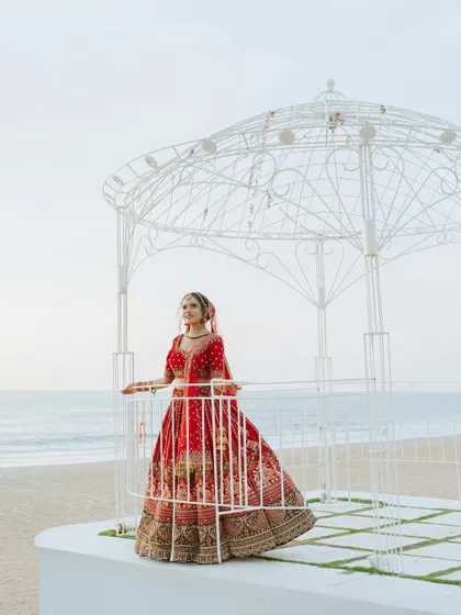 The bride stands in a white gazebo by the sea, her red lehenga a stunning contrast against the serene backdrop.