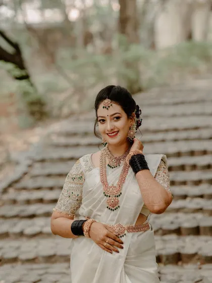 A smiling South Indian bride in a white Kanjeevaram saree, captured outdoors against a natural backdrop. Her happiness radiates in this candid-style portrait.