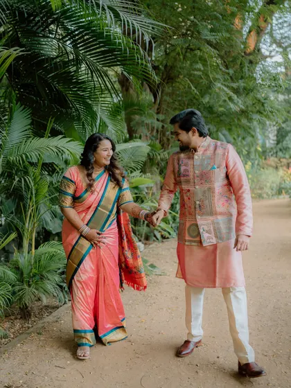 A full-length shot of a couple holding hands and walking along a path in a lush garden. Their coordinated traditional outfits and happy expressions make for a beautiful pre-wedding photo.