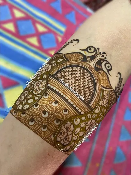A beautiful arm band design featuring two peacocks facing each other. The body of the peacocks is filled with a fine checkerboard pattern, adding texture and detail.