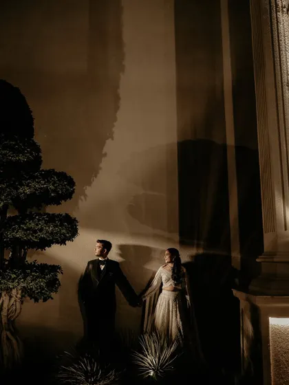 An artistic, low-light portrait of the couple holding hands. The dramatic shadows and architectural elements create a sense of mystery and romance, like a still from a classic film.