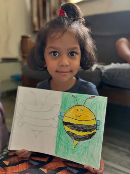A junior KG student holds up her cheerful bee drawing. This exercise helps little ones practice drawing basic shapes and patterns, like the stripes on the bee, while creating a cute character.