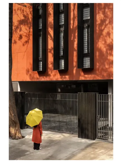 The building's entrance is framed by the strong vertical lines of the security gate and the textured facade. The pop of yellow from the umbrella adds a touch of whimsy to this carefully composed architectural shot.