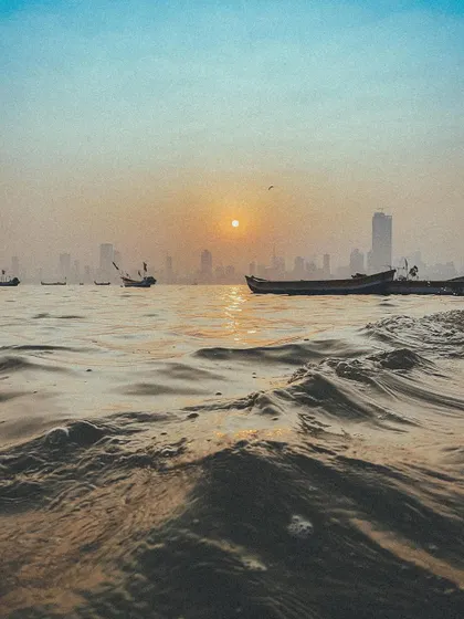 Fishing boats on the water at sunset, with the Mumbai skyline hazy in the background. A peaceful, golden-hour scene.