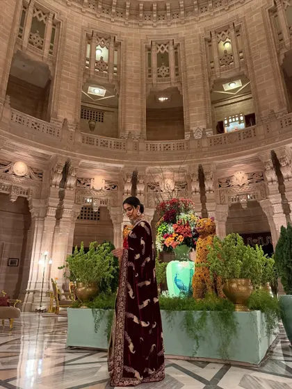 A side profile shot at Umaid Bhawan Palace, showcasing a luxurious maroon velvet saree against the grand, historic architecture.