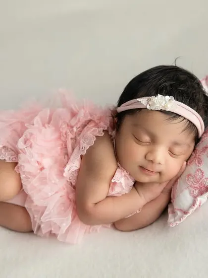 A sweet baby girl in a pink tutu romper, resting her head on a small floral pillow. The white background keeps the image bright and airy.
