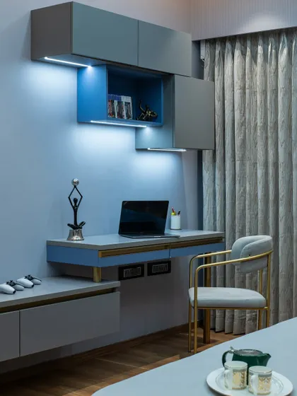 A compact and stylish study corner integrated into a bedroom. The floating desk, overhead cabinets, and task lighting create a functional workspace without taking up too much room. The blue and grey color scheme is calming and conducive to focus.