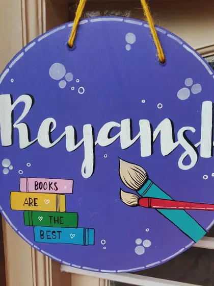 This custom nameplate was designed for a young artist and book lover. It features a stack of books and paintbrushes alongside the name, reflecting the child's passions in a colorful, personalized design.