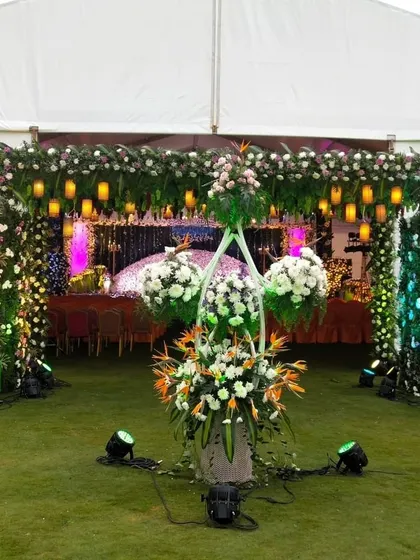 An outdoor event entrance with a floral archway and hanging lanterns, leading into a tented reception area.