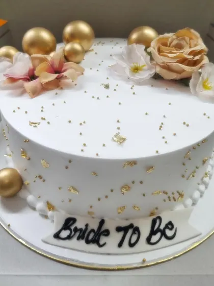 An elegant 'Bride to Be' cake in white, decorated with gold leaf, golden spheres, and delicate flowers. It's the perfect centerpiece for a bridal shower or bachelorette party.