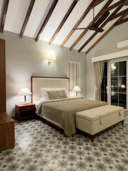 A bedroom in the Chikoo Villa in Goa, showcasing the custom bed with an upholstered headboard and matching storage bench. The patterned floor tiles and exposed ceiling beams add to the room's rustic charm.
