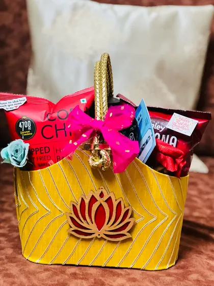 A vibrant yellow fabric basket with a lotus emblem, filled with red-packaged snacks and a bright pink bow.
