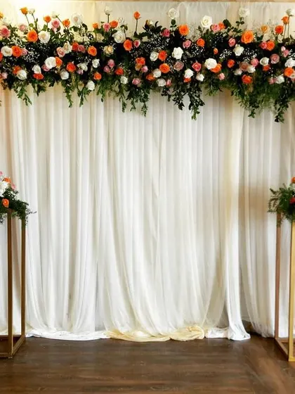 This elegant stage backdrop shows a more contemporary style. A simple white curtain is framed by a lush arch of orange and white roses with cascading greenery, complemented by minimalist gold stands holding matching floral arrangements.