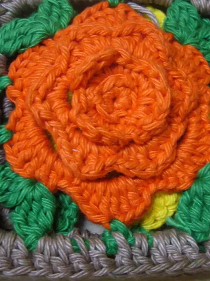 A close-up of a single rose granny square. The three-dimensional orange rose is created by layering petals, with green leaves in the corners.