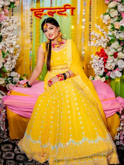 A happy bride poses at her Haldi ceremony. The backdrop is a cheerful mix of yellow drapes, marigold strings, and a large floral arch, perfectly complementing her bright yellow lehenga.