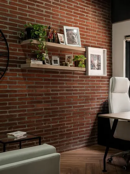 This private cabin uses an exposed brick wall and floating shelves for personal photos and plants to create a warm, personalized, and elegant workspace.