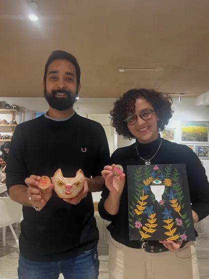A couple poses with their unique creations, a painted cat-themed pot and a dark, floral-inspired canvas.