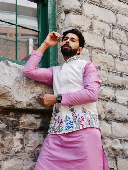 A relaxed pose against a stone wall highlights the contrast between the soft pink kurta and the crisp white of the embroidered Nehru jacket.