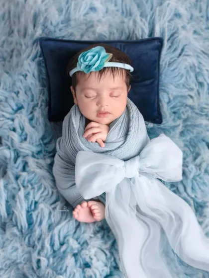 A beautifully swaddled newborn girl sleeping on a soft blue fur rug. The delicate blue floral headband and large bow add a touch of elegance to this peaceful portrait.