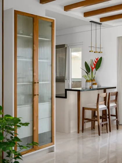 In the 'Malhar' project, the living area overlooks the dining space, which features a full-height crockery unit and a breakfast counter. Exposed ceiling rafters add a charming cottage feel.