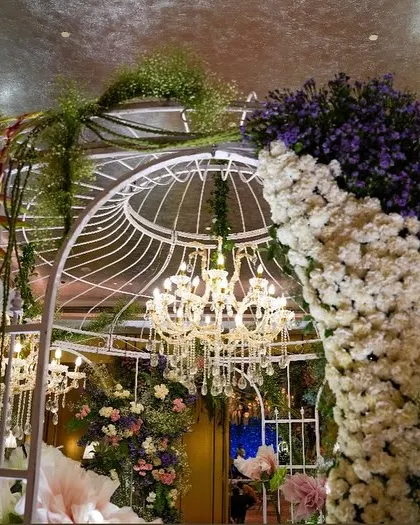 A beautiful white birdcage-style gazebo, adorned with cascading flowers in shades of purple and white. The crystal chandelier inside adds a touch of classic elegance to the whimsical structure.