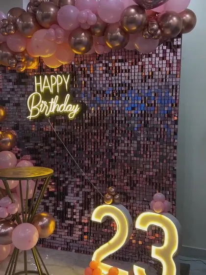 An elegant 23rd birthday setup combining a shimmering rose gold sequin wall with a custom neon sign and large marquee numbers. The organic balloon garland in rose gold and pink tones adds a sophisticated touch, perfect for milestone birthdays and anniversaries.