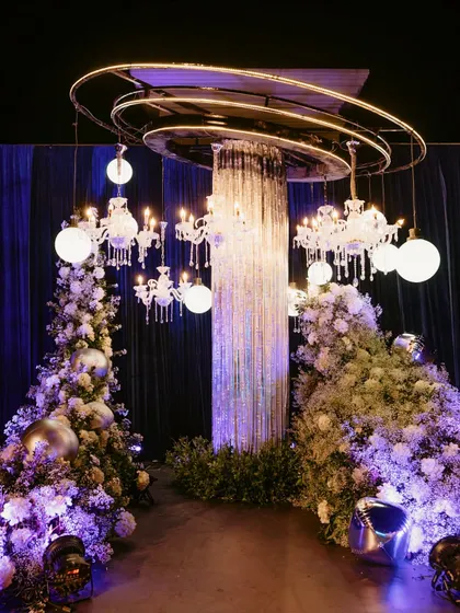 A futuristic entrance installation for a Sangeet, featuring a crystal fringe column, suspended chandeliers, and glowing orbs, all surrounded by clouds of white baby's breath.