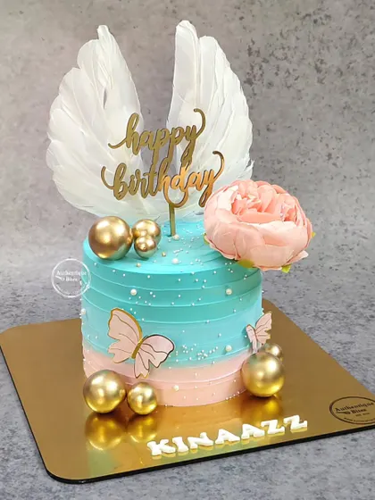 An angelic birthday cake with a tri-color icing effect. The design is topped with delicate white wings, a pink peony, and gold spheres.