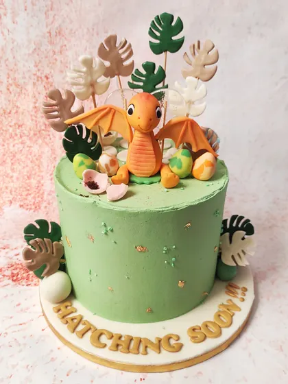 We announce a special arrival with this "Hatching Soon" dinosaur cake. A cute baby pterodactyl sits among its eggs and tropical leaves on a beautiful green cake, making it a unique and adorable theme for a baby shower.