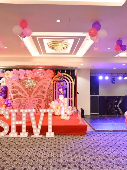 A wide shot of a banquet hall decorated for a butterfly-themed birthday. The view shows the main stage, guest seating, and ceiling balloons, illustrating a complete venue transformation.
