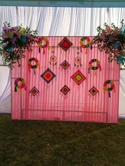 A colorful and rustic backdrop for an outdoor Mehendi function. This setup uses a pink fabric panel decorated with vibrant, handcrafted hangings and is framed by artificial floral arrangements.