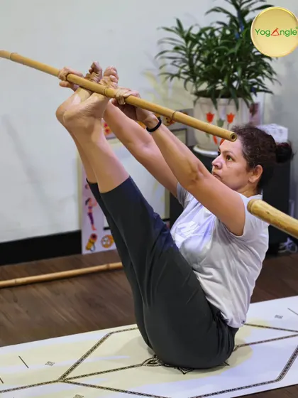 A student uses a bamboo stick to maintain alignment and engage her core in Navasana, or Boat Pose. The stick acts as a guide, ensuring her spine stays straight and her focus remains steady, transforming the pose into an exercise of stability.