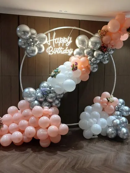A sophisticated birthday backdrop with a circular frame and a balloon garland in pastel pink, peach, and silver. Small floral arrangements are tucked into the balloons for an elegant touch.