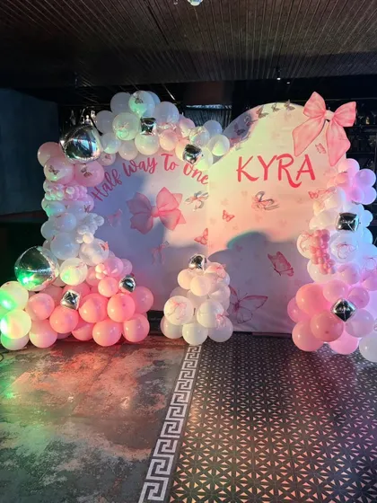 A "Half way to one" celebration for baby Kyra. The backdrop is decorated with a pink and white balloon heart, silver disco balls, and a butterfly and bow theme.