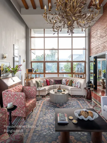 A closer look at the living room in Dhaka, highlighting the custom-made furniture from Cottons and Satins. The curved sofa and patterned armchairs add softness and color, set against a backdrop of exposed brick and a large, expressive area rug.