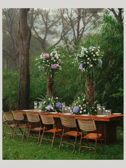 An intimate reception table set for a feast in the heart of nature. The long wooden table, rustic chairs, and towering floral centerpieces create an elegant yet grounded atmosphere.