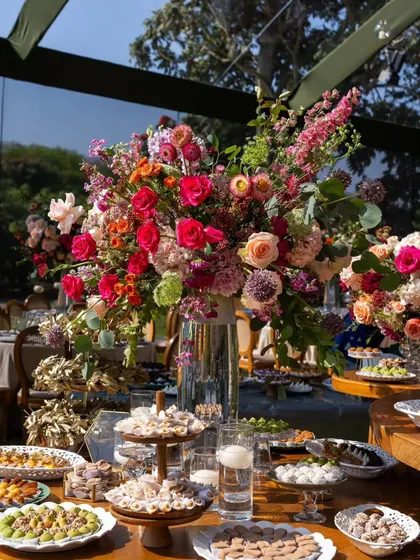 A lavish dessert and sweets table featuring an explosion of colorful flowers in a large glass vase. The abundance of both sweets and florals creates a feeling of luxurious celebration.