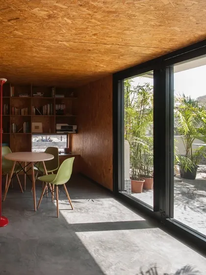 A compact workspace designed for a journalist, featuring floor-to-ceiling glass and warm OSB wood panels. The design maximizes natural light and connects the interior to an outdoor terrace, making a small space feel open and integrated with nature.