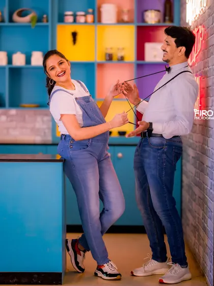 A candid, laughing moment from the same casual maternity shoot. The colorful kitchen-themed set provides a fun, quirky backdrop as the mom-to-be playfully tugs on her partner's suspenders.